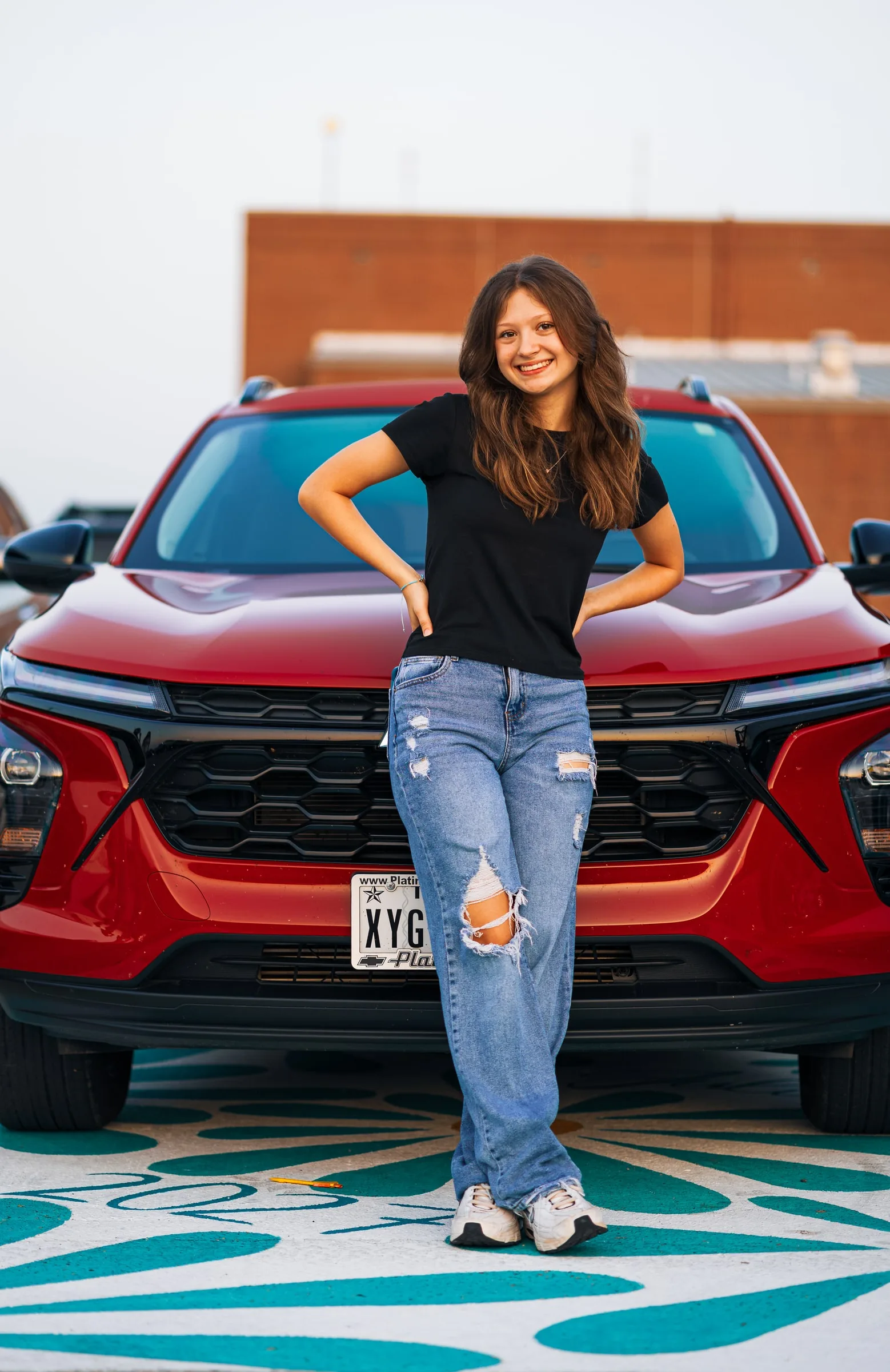 Senior portrait of a student standing in front of a red SUV, hands on hips
